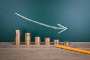 Stacks of coins in descending order with an arrow drawn on the wall indicating a downward trend, accompanied by a yellow pencil.