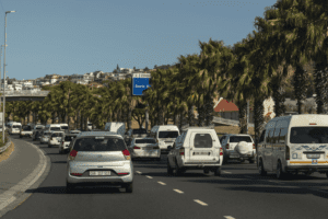 Traffic on a palm-lined road with clear skies and a hillside residential area in the distance.