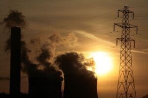 Silhouetted industrial power plant with cooling towers and smokestack emissions against a sunset, with electricity pylons in the foreground.