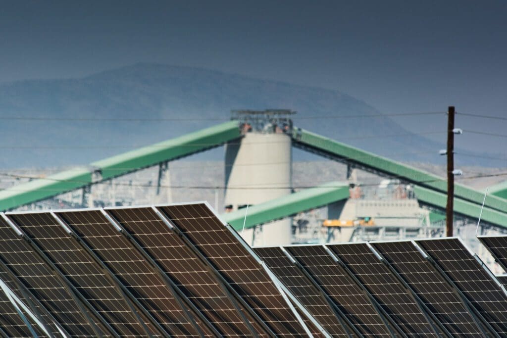 Solar panels in the foreground with industrial equipment in the background against a hazy sky.