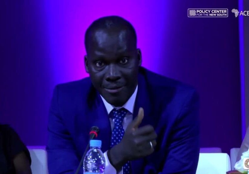 A man in a dark suit speaking at a panel discussion with a microphone in front of him.