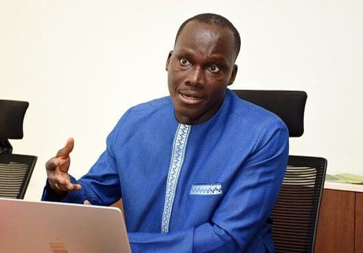 A man in a blue traditional attire sitting behind a laptop and gesturing during a conversation.