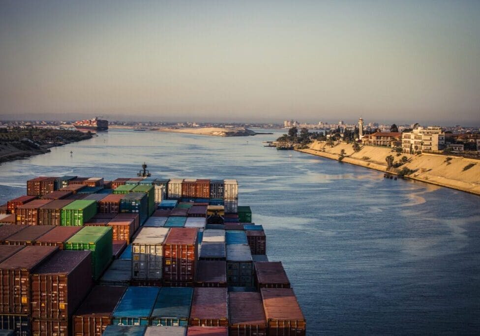 Cargo ship loaded with containers navigating through a wide river with a coastal cityscape in the background at dusk.