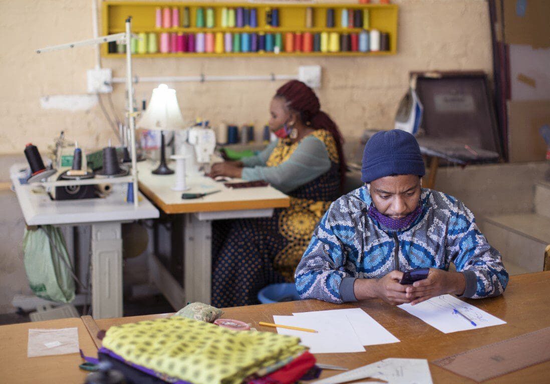Two individuals focused on their tasks in a tailoring workshop, with sewing machines and colorful thread spools in the background.