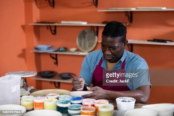 Man painting ceramics in a workshop surrounded by colorful paints.