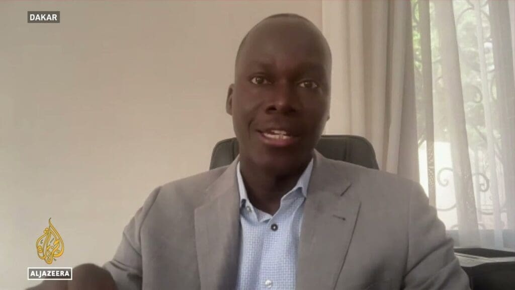 A man in a gray suit jacket speaking during a video interview with al jazeera, with "dakar" indicated as the location.