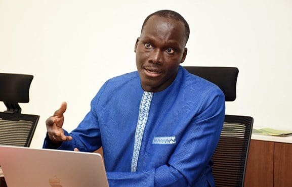 A man in a blue traditional attire sitting behind a laptop and gesturing during a conversation.