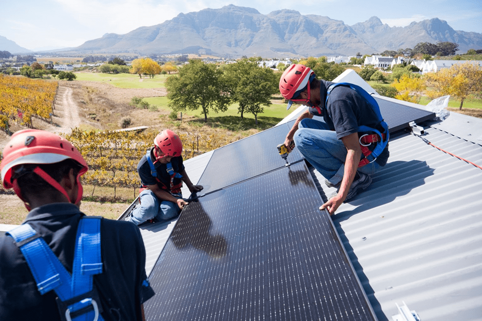 Workers installing solar panels on a roof with a mountainous landscape in the background.