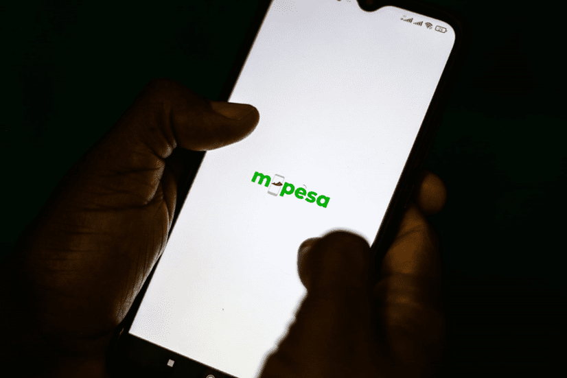 A person holding a smartphone displaying the m-pesa logo on the screen against a dark background.