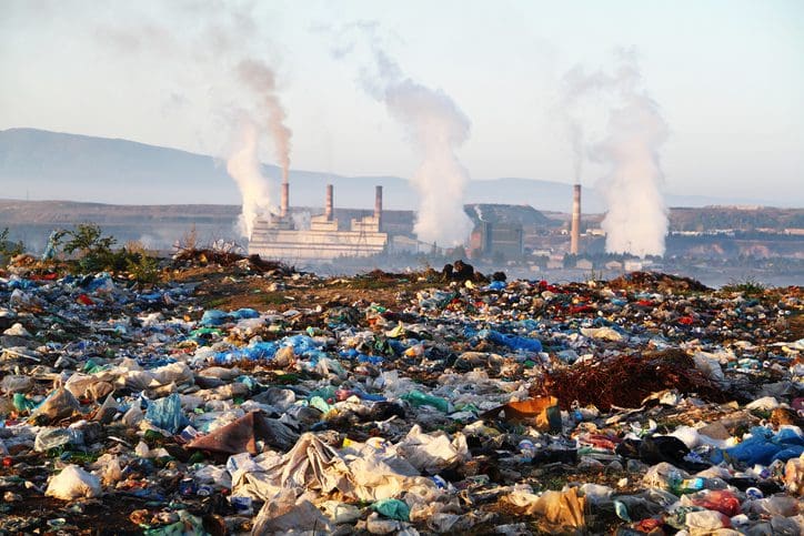 A landfill in the foreground with industrial smokestacks emitting smoke in the background.