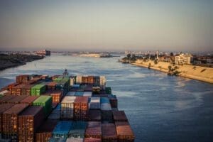 Cargo ship loaded with containers navigating through a wide river with a coastal cityscape in the background at dusk.