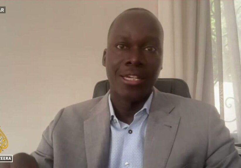 A man in a gray suit jacket speaking during a video interview with al jazeera, with "dakar" indicated as the location.