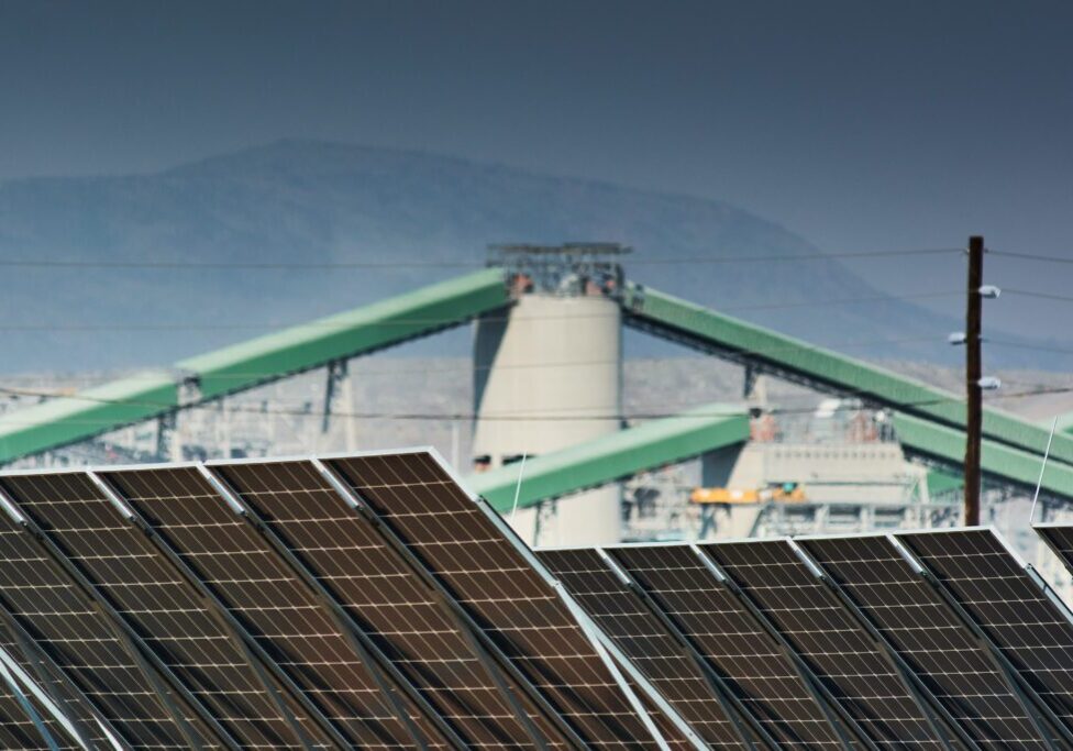 Solar panels in the foreground with industrial equipment in the background against a hazy sky.