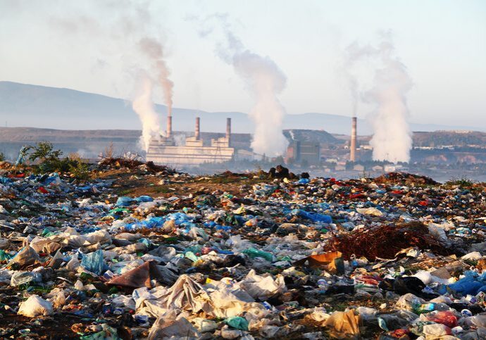 A landfill in the foreground with industrial smokestacks emitting smoke in the background.
