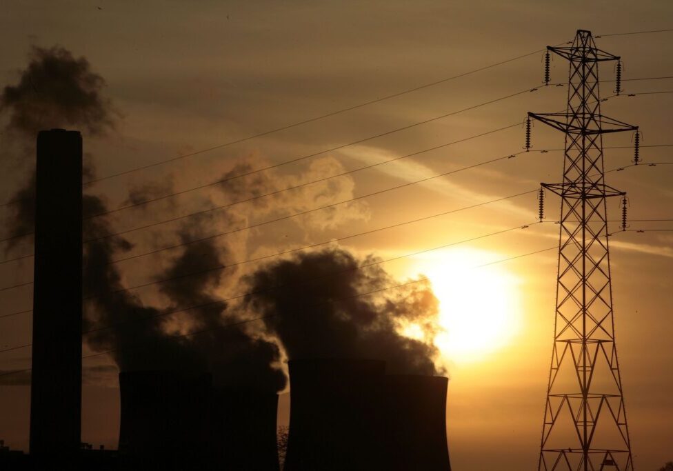 Silhouetted industrial power plant with cooling towers and smokestack emissions against a sunset, with electricity pylons in the foreground.